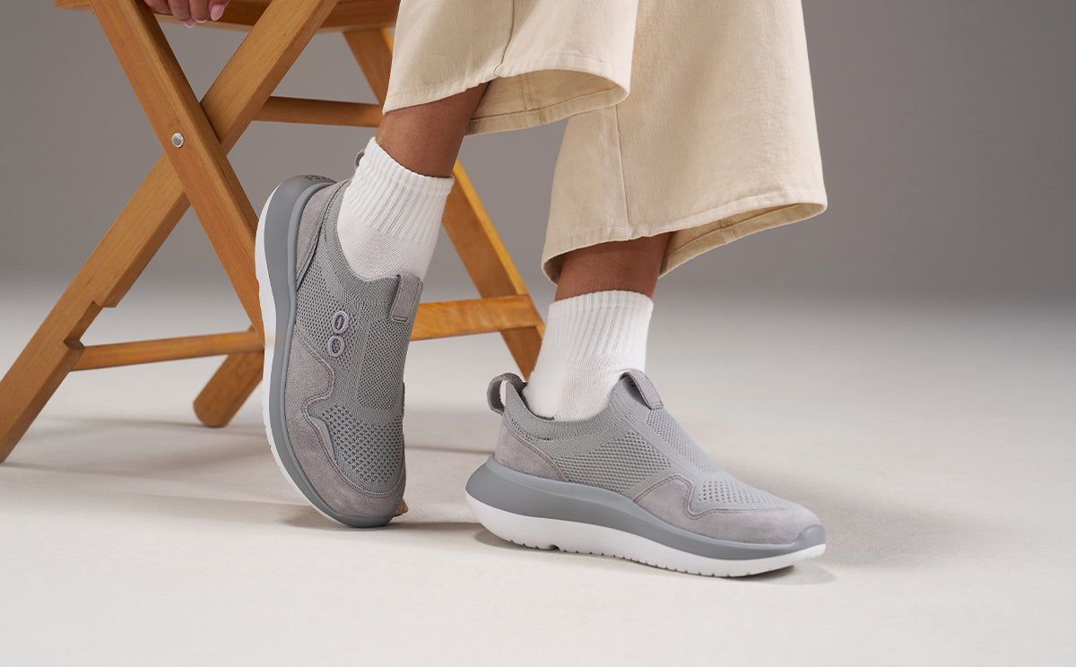 A person wearing light gray slip-on sneakers with white crew socks and beige trousers, seated on a wooden folding chair against a neutral studio background | lifestyle