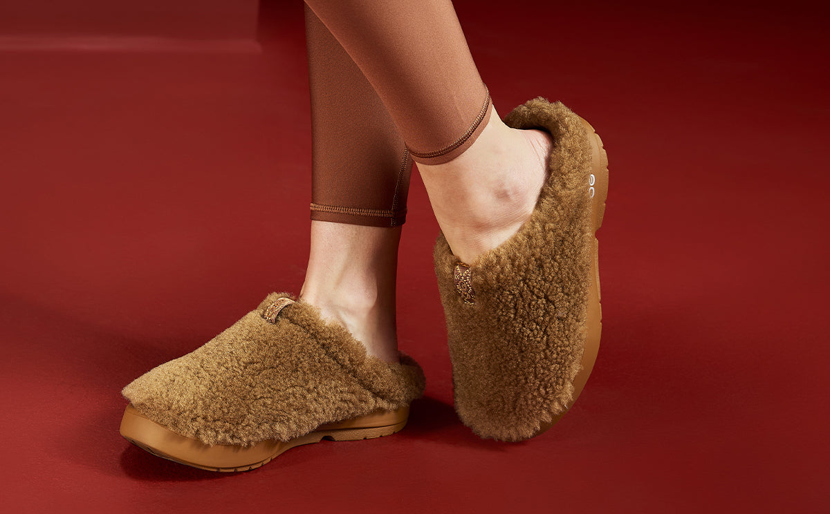 A person wearing fluffy tan shearling slip-on slippers with tan soles, standing against a solid dark red background | lifestyle