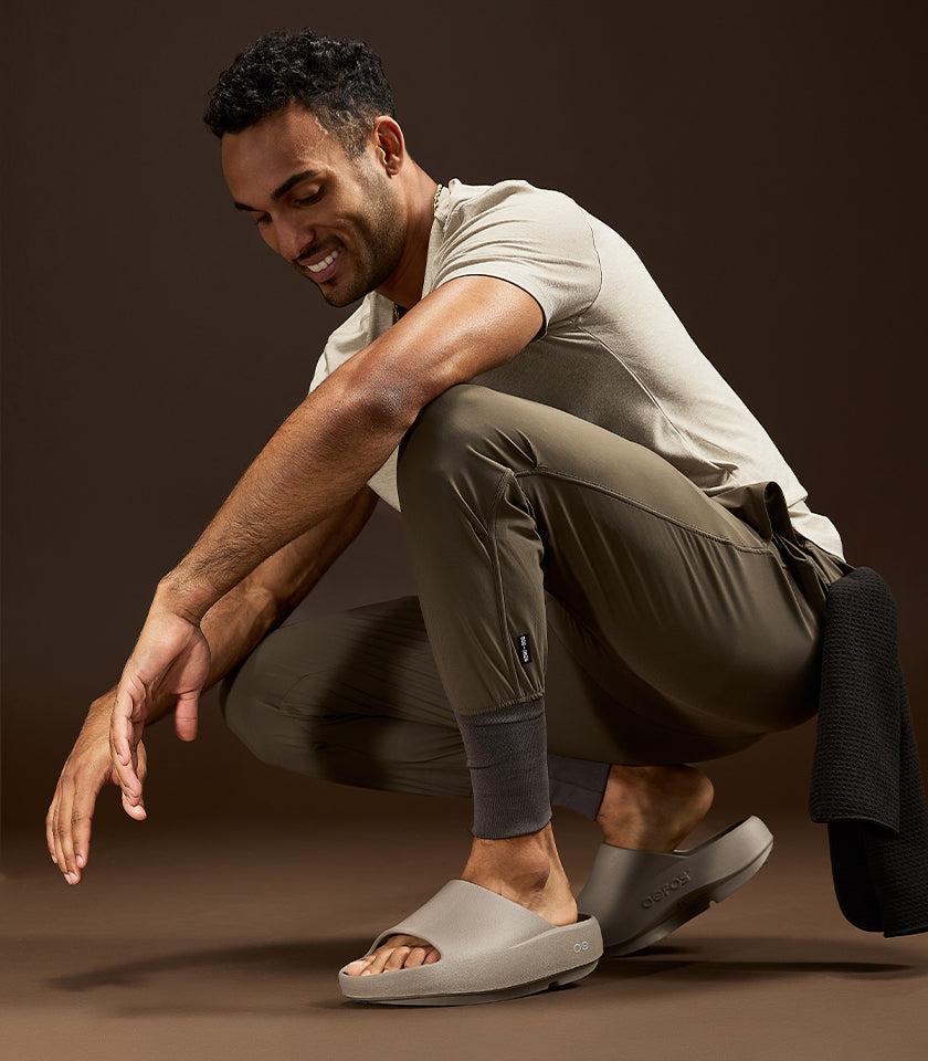 A man crouches in a studio wearing a light beige t-shirt, olive green athletic pants, and matching gray slide sandals. A black knit towel is draped over his leg against a solid dark brown background.