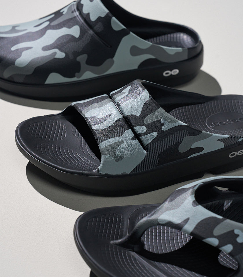 Three black slide sandals with gray and black camouflage straps, arranged diagonally on a light gray surface.