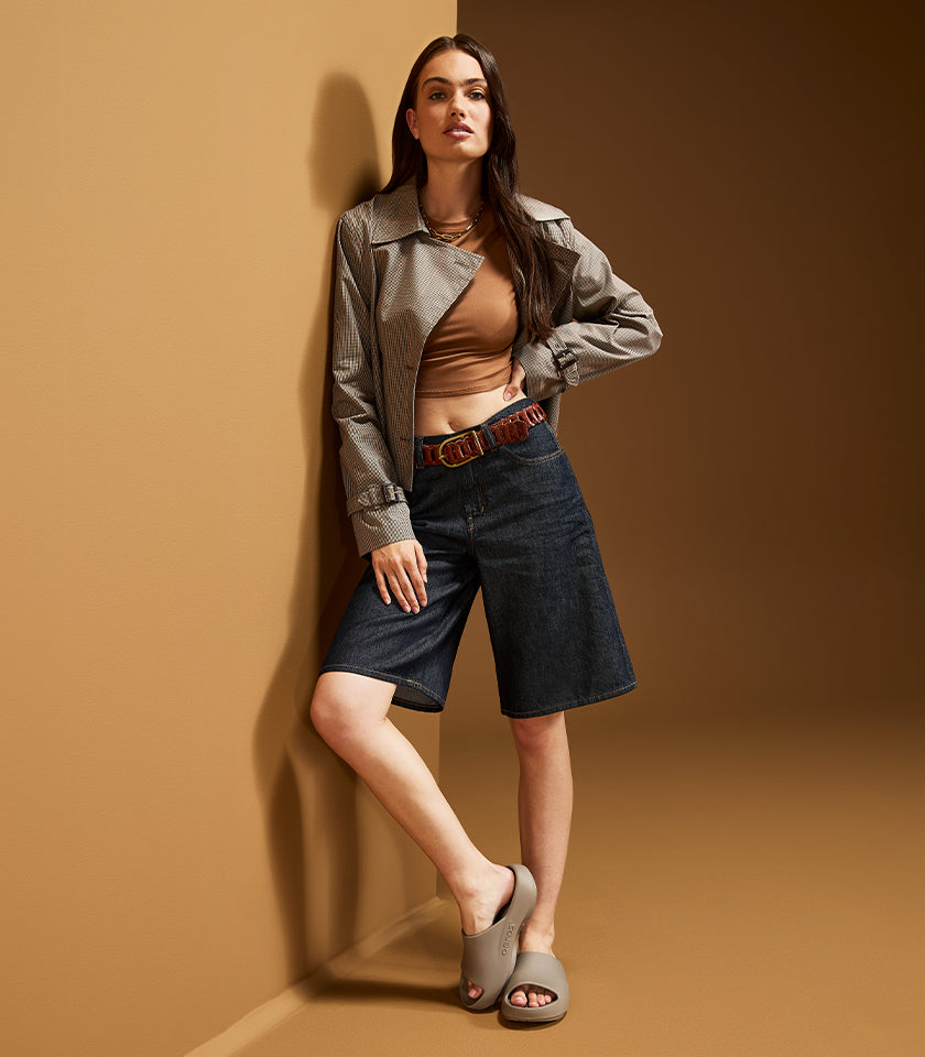 A model leans against a warm brown studio wall wearing a gray cropped jacket over a tan crop top, dark blue denim shorts, and gray slide sandals.