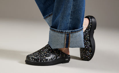 A person wearing black clogs with a multicolor speckled pattern and blue jeans, shown from the calves down against a neutral studio background | lifestyle