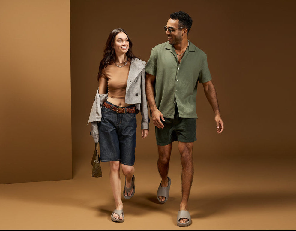 A man and a woman walking side by side in a studio setting with a warm brown background. The man wears a muted green short-sleeve button-up shirt, dark green shorts, and gray slide sandals.