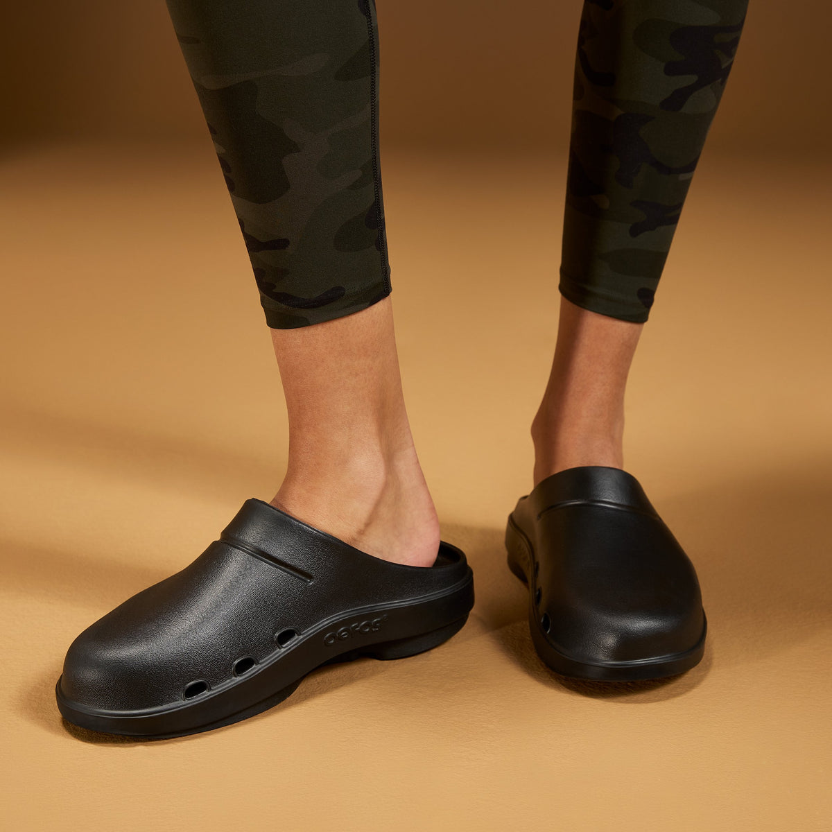 Black slip-on clog-style shoes worn with dark camouflage leggings, shown on a model against a warm tan studio background.