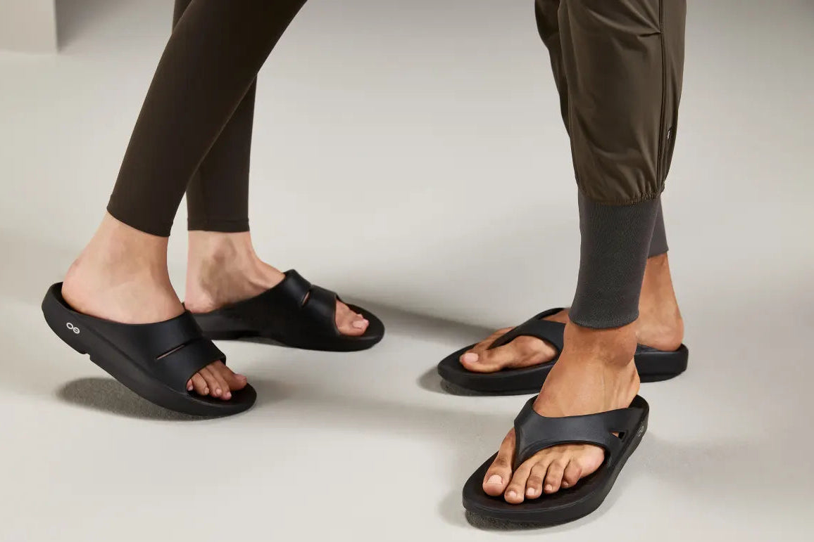 Black slide sandals worn by a model, shown from the feet up in a clean studio setting with a light gray floor and background.