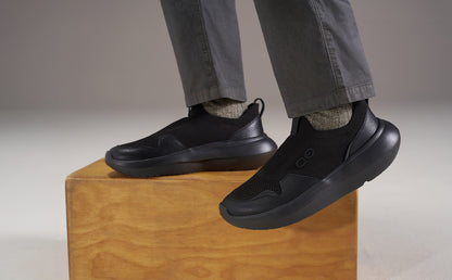 A person wearing black slip-on sneakers with gray pants and socks, standing on a wooden block against a neutral studio background | lifestyle