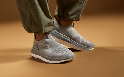 A person wearing light gray slip-on sneakers and olive green pants, standing on a warm brown studio floor | ifestyle