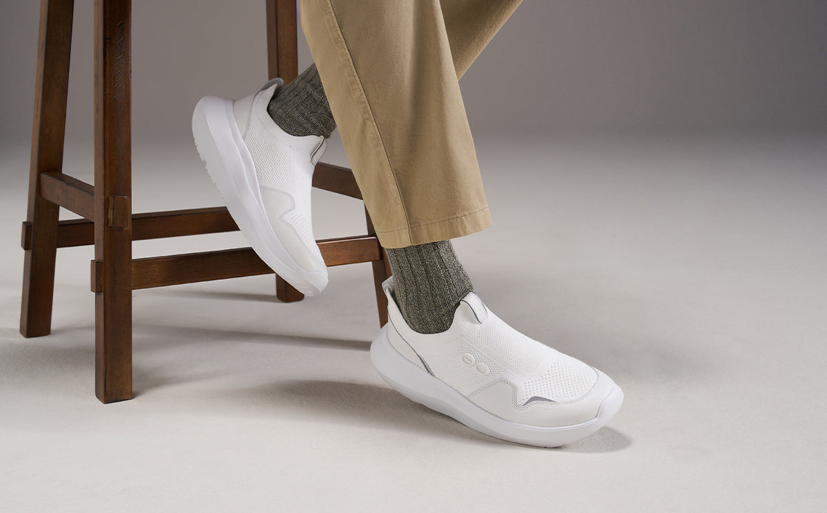 White slip-on sneakers worn with gray socks and beige trousers, shown on a person seated on a wooden stool against a neutral studio background | lifestyle