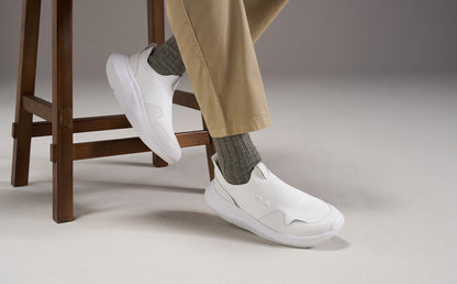 White slip-on sneakers worn with gray socks and beige trousers, shown on a person seated on a wooden stool against a neutral studio background | lifestyle