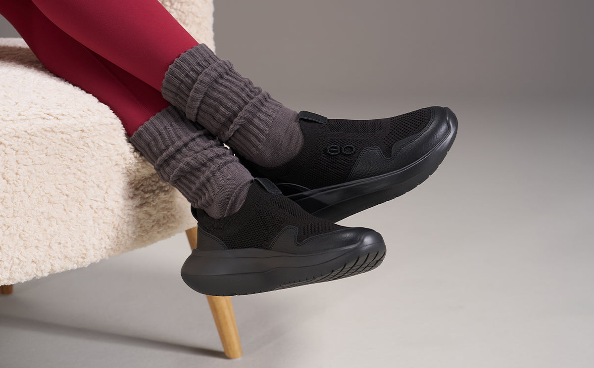 A person seated on a cream chair wearing black slip-on shoes with gray ribbed socks, against a neutral studio background | lifestyle