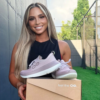 A woman stands outdoors holding a pair of light purple sneakers resting on a brown shoebox with the brand name 'feel the oo.' printed on it.