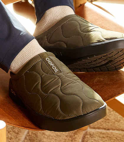 Green quilted slippers with Oofos branding worn indoors on a wooden floor.