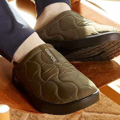 Green quilted slippers with a brand logo worn indoors on a wooden stool.