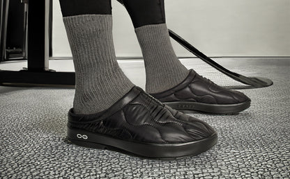 Men's OOcoozie Thermo Mule - Black model standing inside wearing shoes with hockey stick next to him | lifestyle
