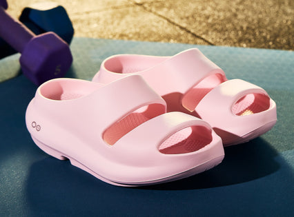 A pair of light pink molded slide sandals with wide double straps, displayed on a blue surface with a purple dumbbell in the background.