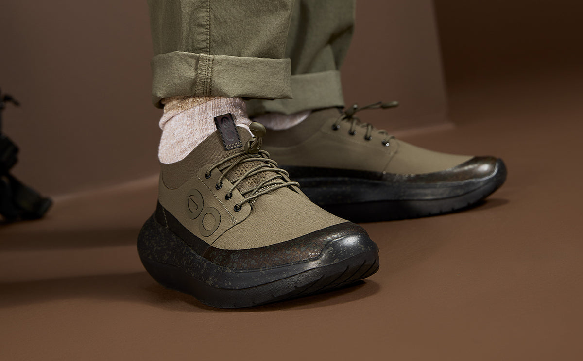 A close-up of a person wearing olive green low-top lace-up shoes with chunky black soles, paired with light pink socks and cuffed olive pants, set against a warm brown studio background | lifestyle