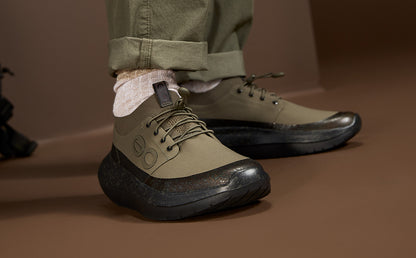 A close-up of a person wearing olive green low-top lace-up shoes with chunky black soles, paired with light pink socks and cuffed olive pants, set against a warm brown studio background | lifestyle