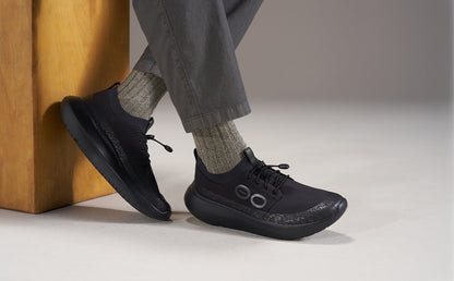 A person wearing black athletic sneakers with gray socks and gray pants, seated on a wooden block against a neutral studio background | lifestyle