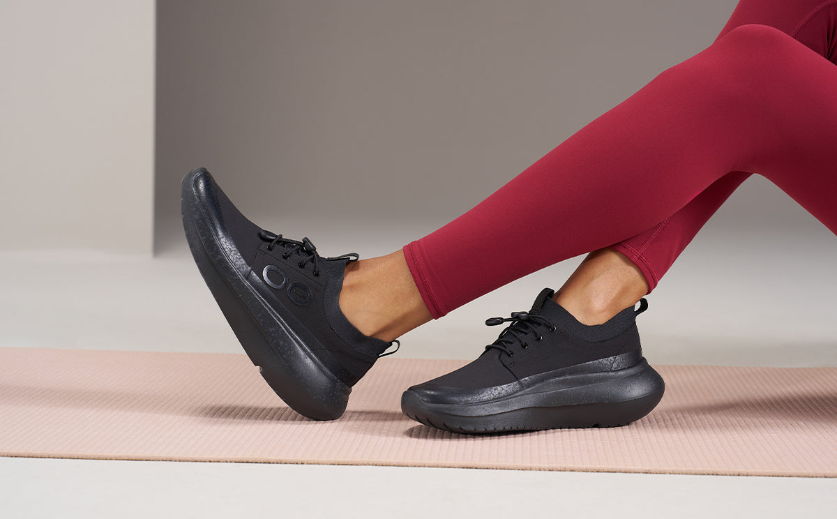 A person wearing black athletic sneakers and red leggings sitting on a pink yoga mat in a minimalist studio setting | lifestyle