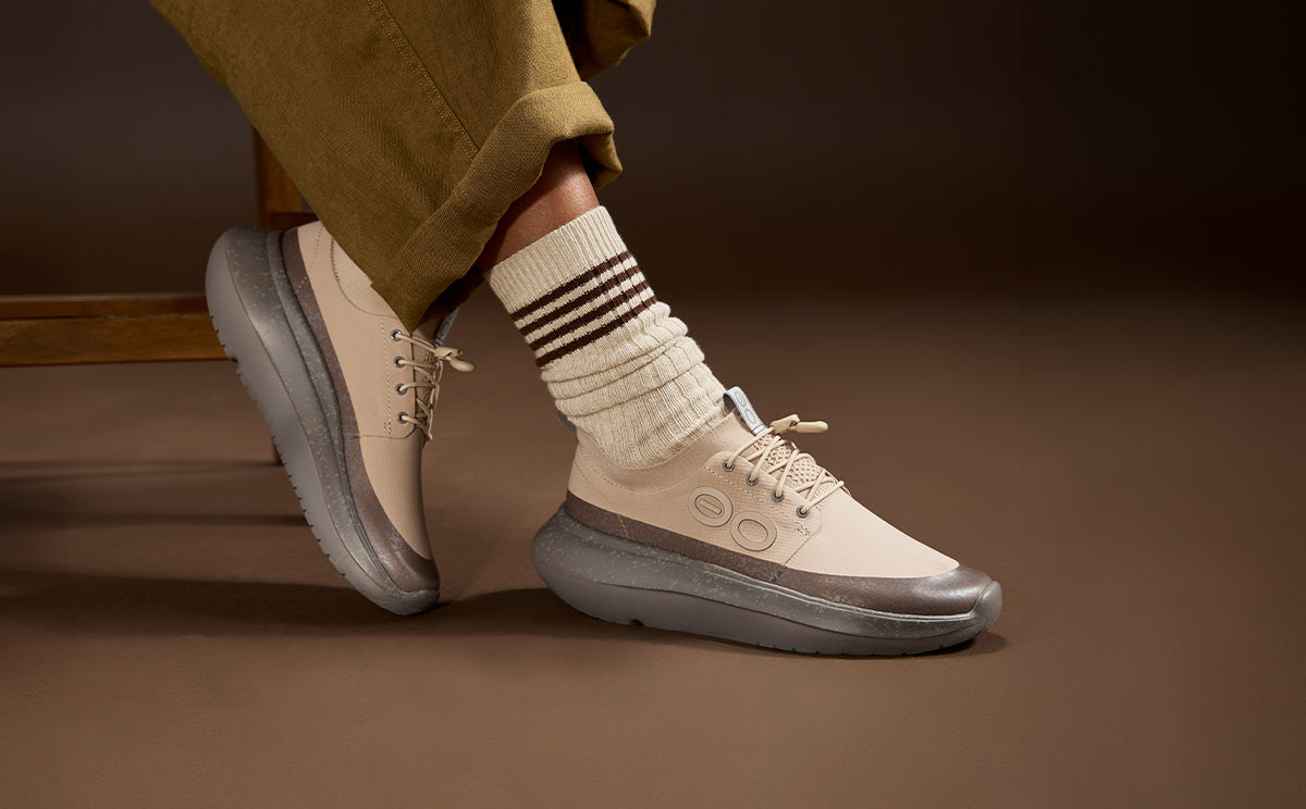 A person wearing beige and gray platform sneakers with cream socks, shown from the knee down in a studio setting with a warm brown background | lifestyle