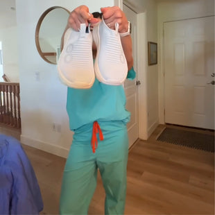 Person in teal scrubs holding white shoes in a room with wooden flooring and a door.