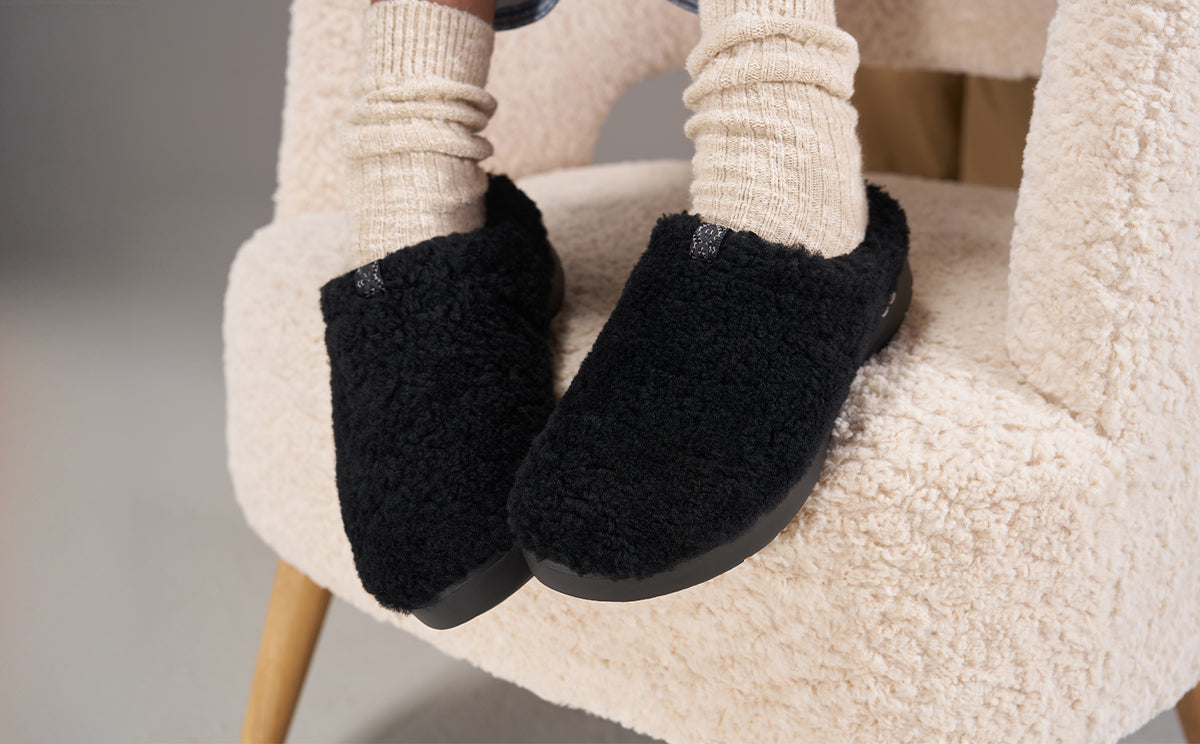 A close-up of a person sitting on a cream-colored sheepskin chair, wearing black fuzzy slip-on slippers and cream-colored socks | lifestyle