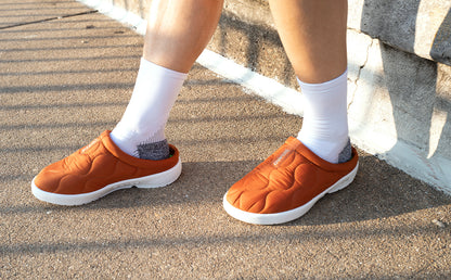 A person wearing orange slip-on shoes with white socks, standing on a paved outdoor surface next to a stone wall | lifestyle