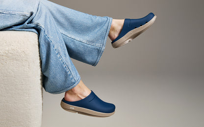 A person wearing blue clogs with light beige soles, sitting on a cream-colored chair. The person is also wearing light blue jeans, set against a neutral gray studio background | lifestyle