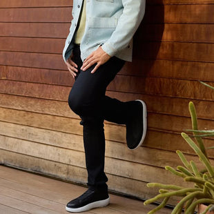Person wearing black pants and shoes leaning against a wooden wall with a plant in the foreground.
