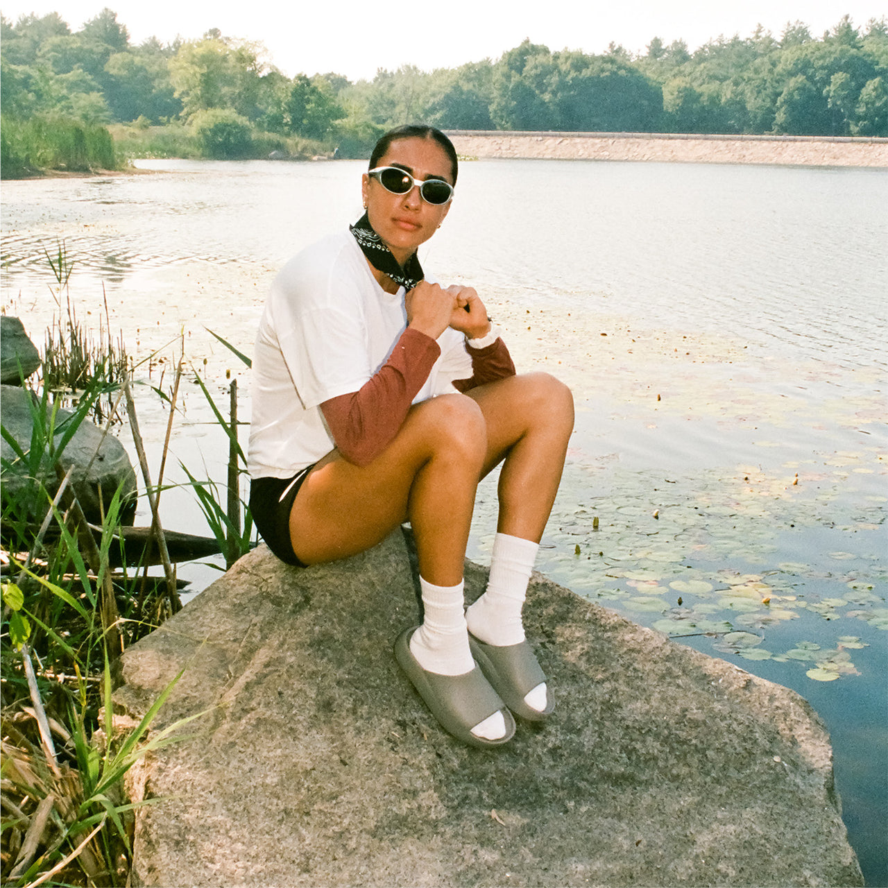 A person sitting on a large rock by a lake, wearing a white t-shirt, brown long-sleeve top, black shorts, white socks, gray slip-on shoes, and sunglasses.