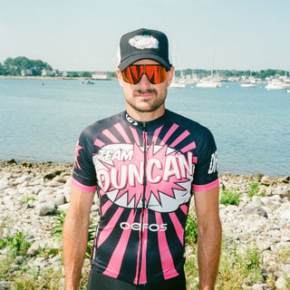 A man wearing a black and pink cycling jersey with 'Team Duncan' and 'Geefos' branding, black cycling shorts, and a black cap stands outdoors near a rocky shoreline with water and boats in the background.