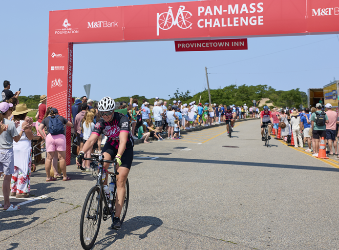 A cyclist rides through a race finish line under a large red 'Pan-Mass Challenge' arch, with spectators lining the road and other riders in the background.
