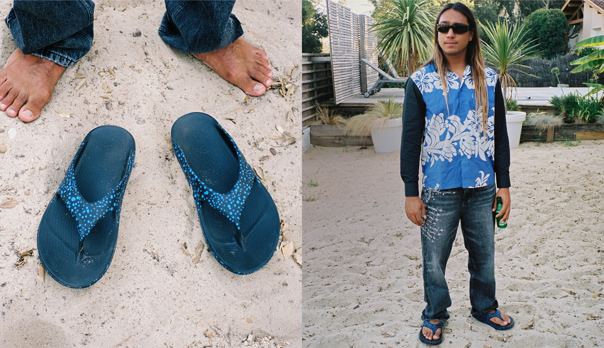A split image showing a pair of dark blue patterned thong sandals on sand next to a person wearing them outdoors. The sandals have a dark blue base with a lighter blue and white patterned strap.