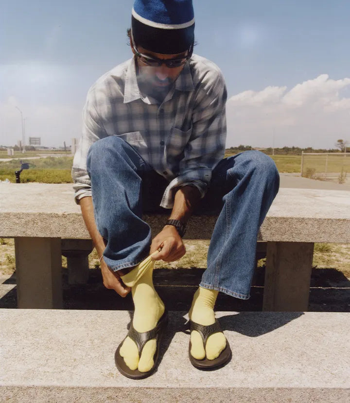 A man sitting on a concrete bench outdoors wearing a blue and white plaid shirt, blue jeans, a blue and white beanie, sunglasses, and yellow socks with OOFOS × Adsum OOriginal Limited Sandals.