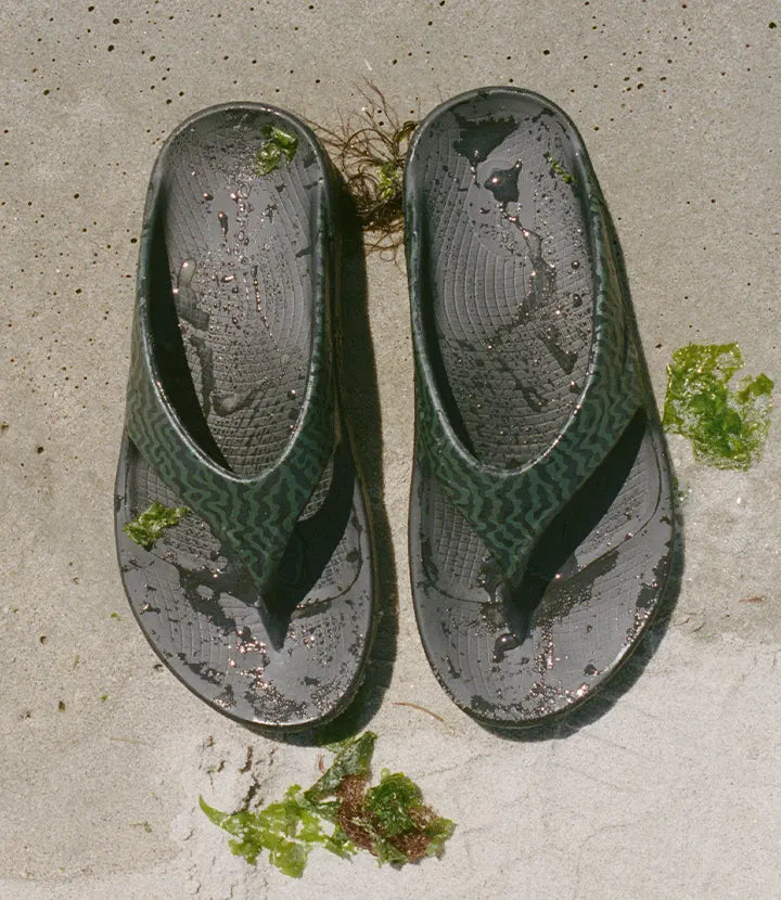 A pair of OOFOS × Adsum OOriginal Limited Sandals with green seaweed on a sandy beach.