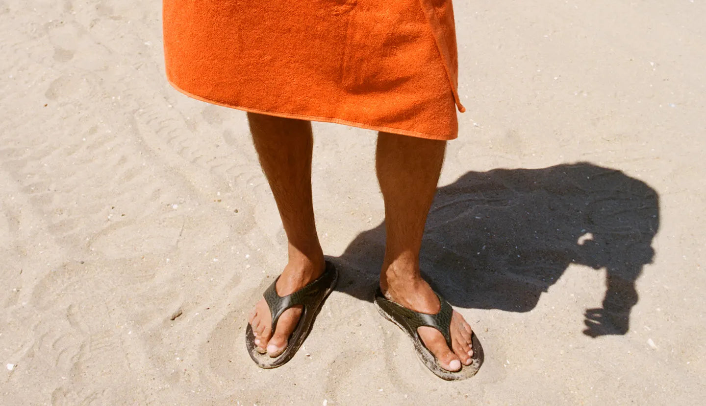 An orange beach towel wrapped around the lower half of a masculine person standing on sand, wearing OOFOS × Adsum OOriginal Limited Sandals.